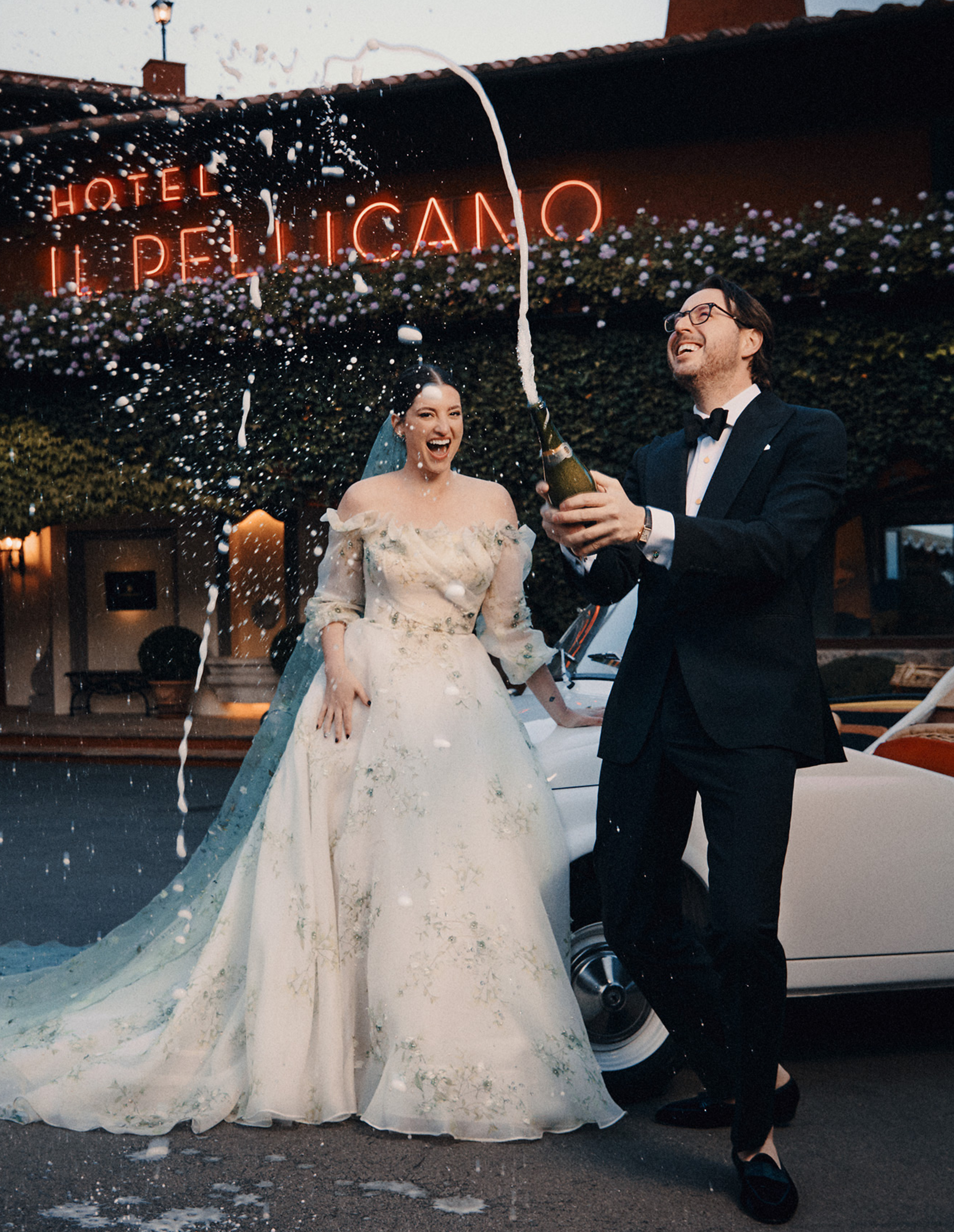 Newlywed couple popping a bottle of champagne in front of Hotel Il Pellicano.